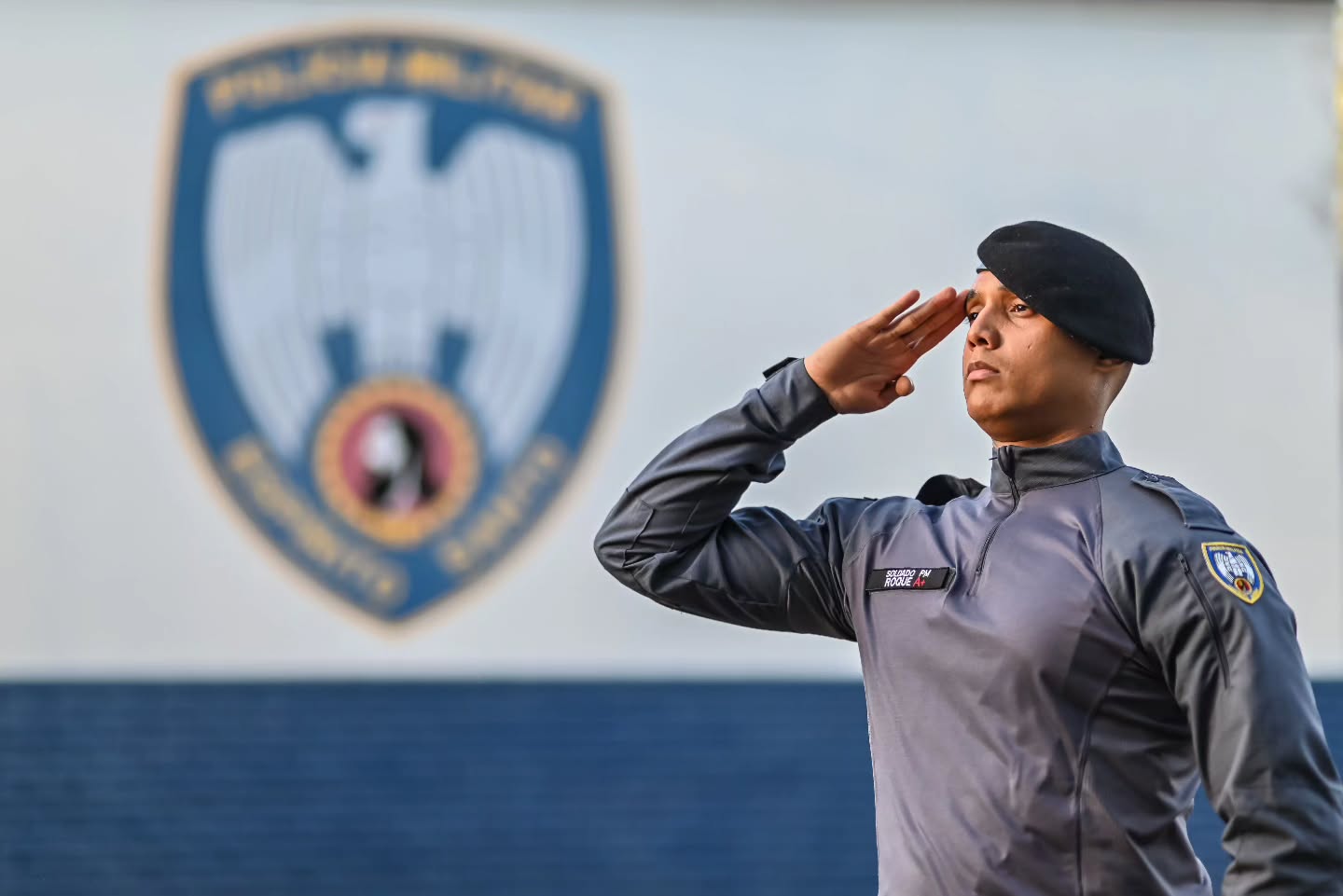 Soldado da Polícia Militar do Espírito Santo em continência diante do brasão institucional da PMES — símbolo da regra editalícia que a Justiça do Estado já analisou em casos de eliminação por falta de CNH